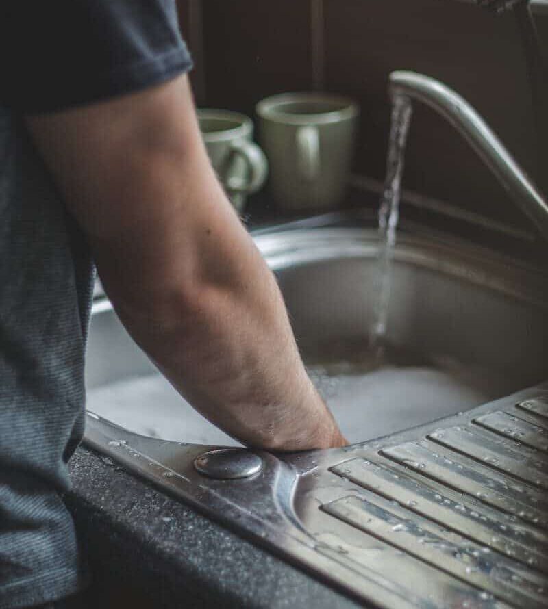A man washing his hands at a kitchen sink with cups and cleaning products nearby.